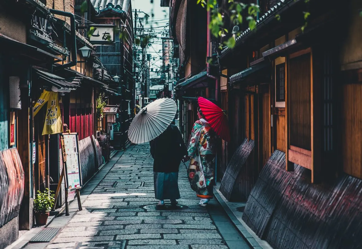 Two people walking down a narrow alley with umbrellas