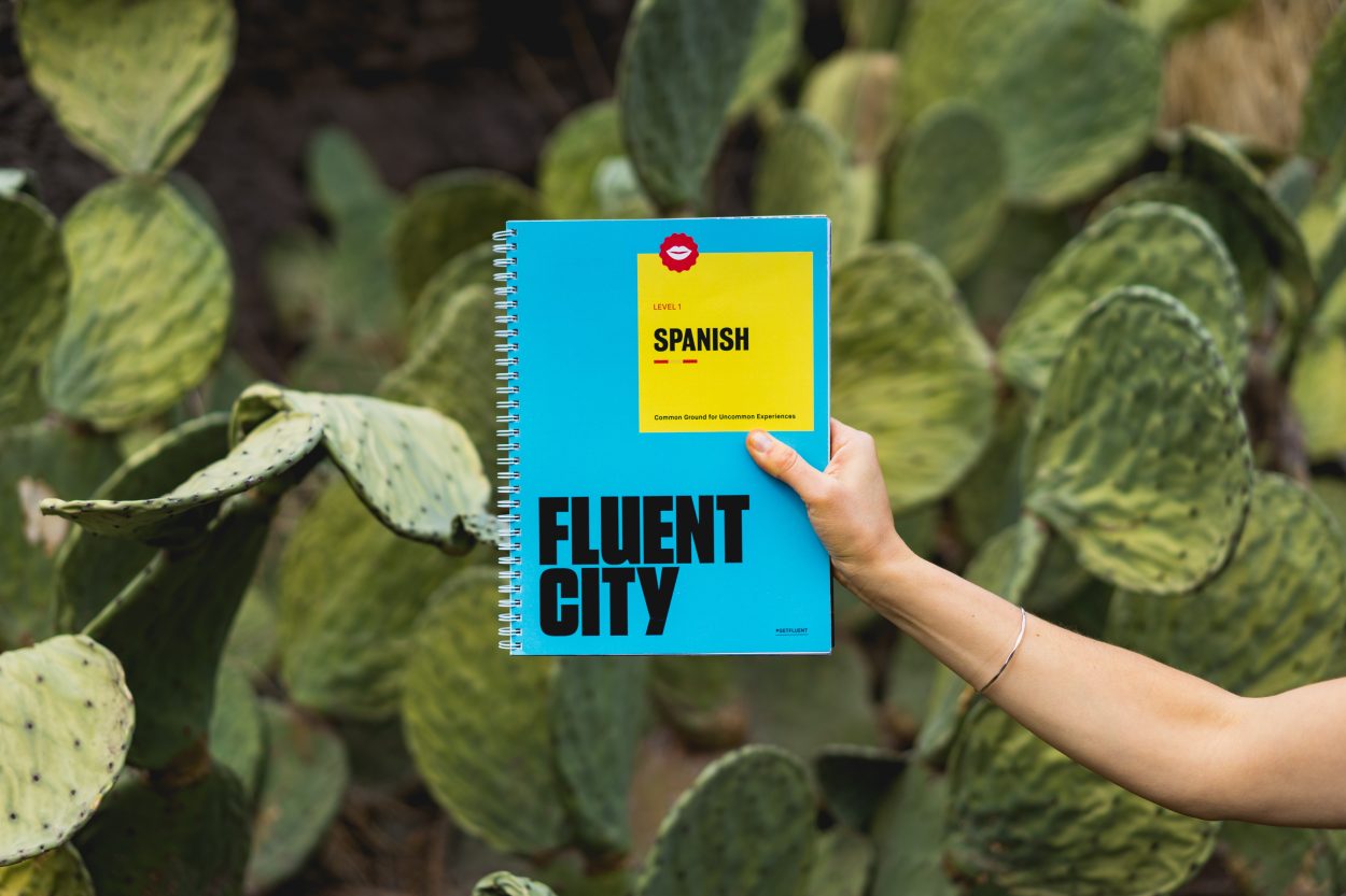 An arm of a person with a book in her hand which says "Spanish Fluent City" with a bunch of plants in the background.