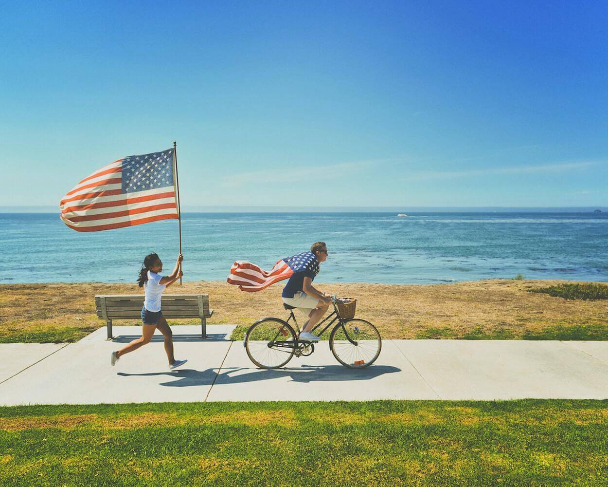 A boy riding his bicycle with an American flag on his shoulders. A girl holding an American flag while running.