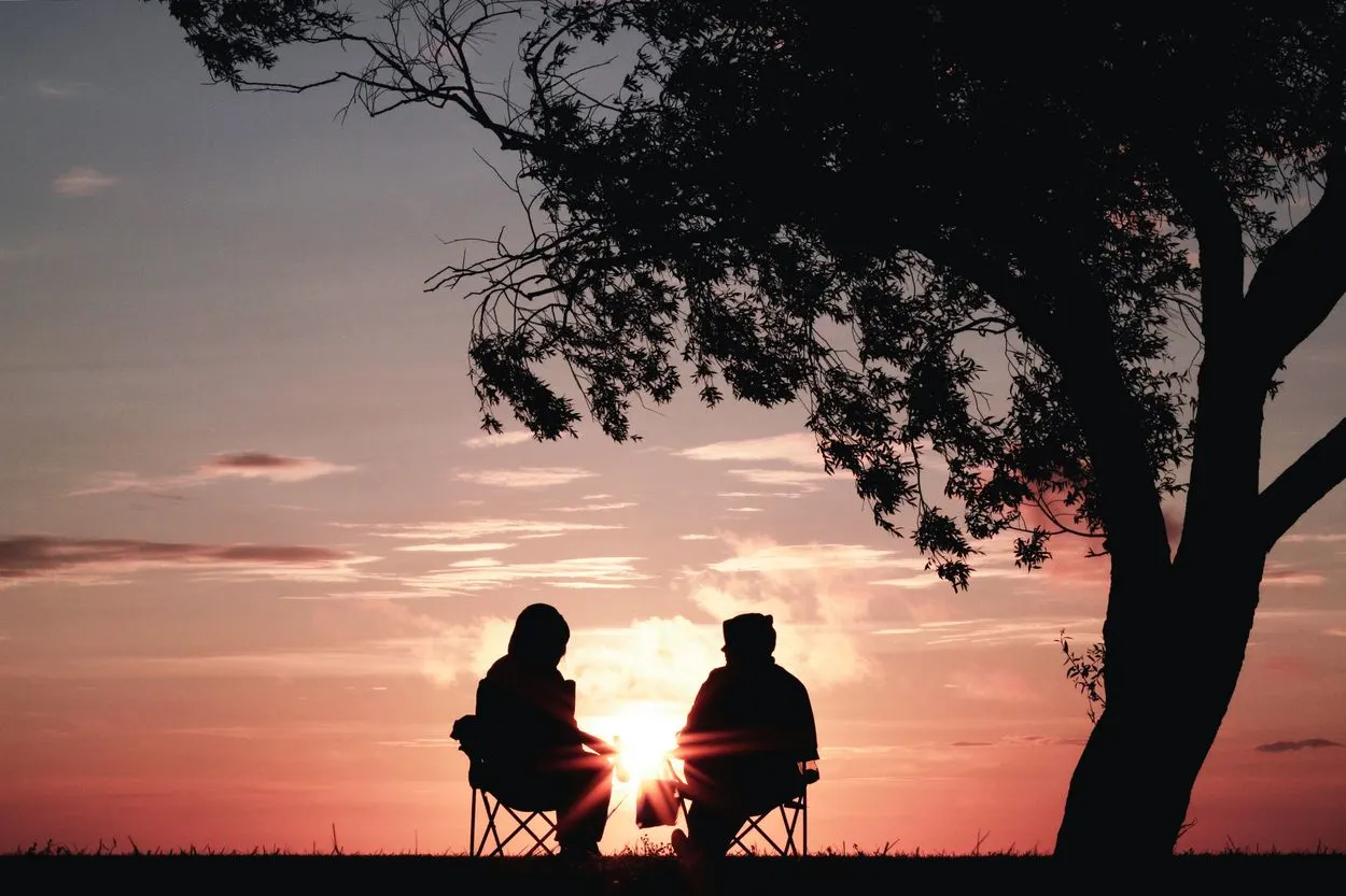 A silhouette of two people sitting next to a tree in front of a sunset