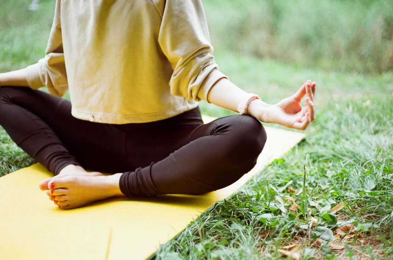 A woman sitting on the yoga mat which is on the grass doing yoga in a black tights and green top