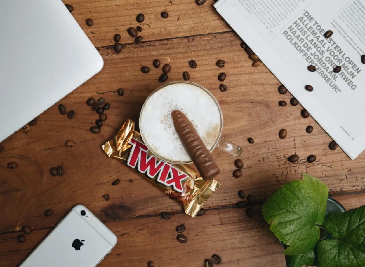 A cup of coffee with an unwrapped Twix bar inside it and a wrapped one placed beside it.