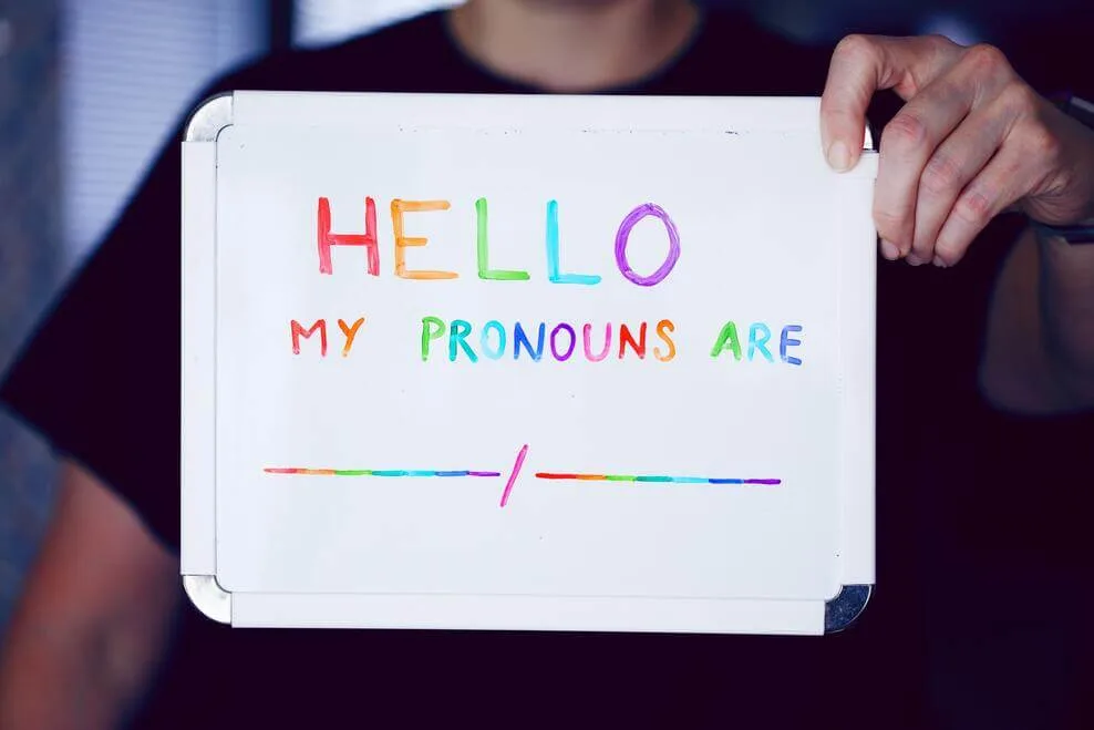 A photograph showing a man holding a flashcard with "Hello" "my pronouns are" written on it