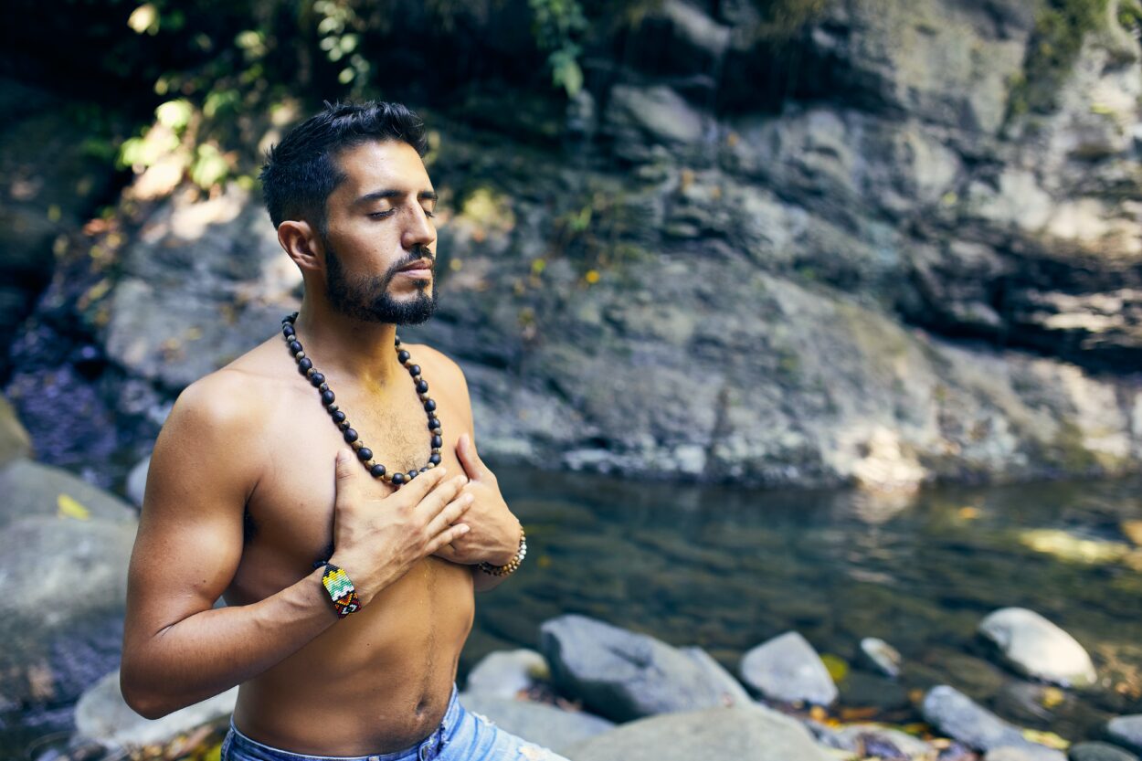 Topless man meditating near water.

